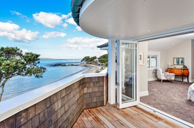 A bright coastal balcony or terrace overlooking a sheltered bay, with timber decking, a shingle-clad balustrade, and an open glass door leading into a softly furnished bedroom or sitting area. The curved white soffit and sweeping glazing create a contemporary seaside architectural feel.