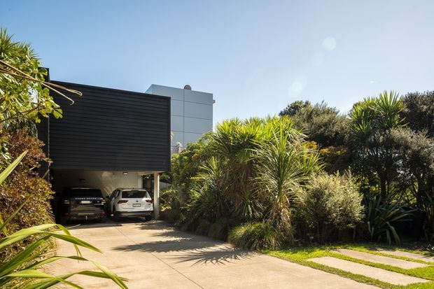 Exterior view of a contemporary residential property with a dark horizontal-clad carport or garage volume, a light rendered upper structure, and a landscaped driveway bordered by dense ornamental grasses and spiky coastal-style planting.