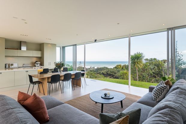 Bright open-plan contemporary living and kitchen area with a large dining island, extensive floor-to-ceiling glazing, and views over landscaped greenery toward the ocean. Neutral built-in cabinetry, grey sectional sofas, timber flooring, and a round coffee table create a relaxed coastal setting.