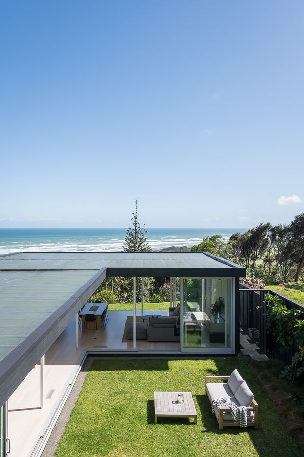 A modern low-profile coastal house arranged around a small central grass courtyard. The image looks over a flat metal roof and extensive floor-to-ceiling glazing toward a visible ocean shoreline, with a minimalist outdoor seating area and compact interior living and dining spaces beneath a covered roof.