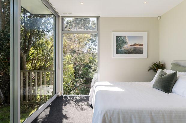 Bright, serene bedroom with a neatly made bed, soft neutral bedding, dark carpet, and pale walls. Floor-to-ceiling glazed doors and windows open to leafy garden views, while a framed coastal landscape artwork and small floral arrangement add understated decoration.