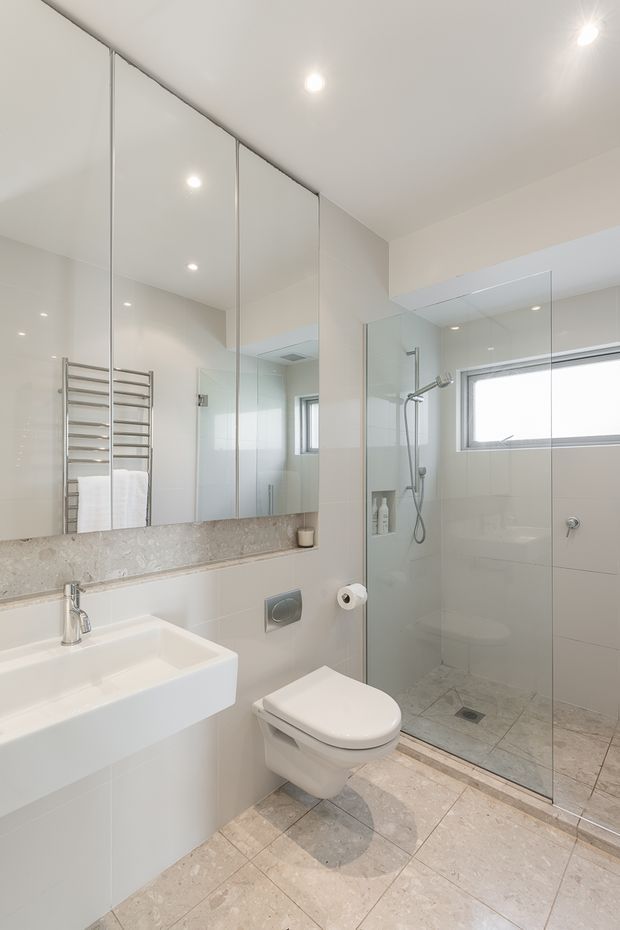 Bright contemporary bathroom with a wall-mounted white vanity basin, concealed-cistern toilet, frameless glass walk-in shower, large mirrored wall cabinetry, chrome heated towel rail, pale stone-look tiled floor, and a narrow horizontal window. The room has a clean, neutral finish with recessed ceiling lights.