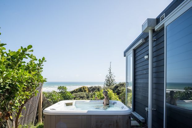 Exterior view of a modern coastal home with dark horizontal cladding and large reflective glazing, opening onto a private spa pool. Ocean waves and landscaped planting are visible beyond the property, creating a sheltered outdoor bathing area.