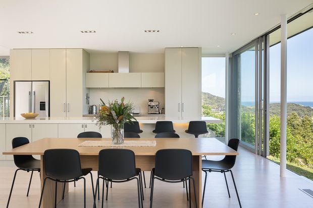 Bright open-plan kitchen and dining area with extensive cream built-in cabinetry, integrated appliances, a long timber dining table with black dining chairs, and full-height glazed sliding doors opening to a leafy coastal landscape. The room has a clean contemporary feel and abundant natural light.