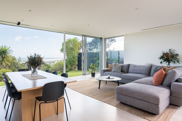Bright, open-plan contemporary living and dining area with floor-to-ceiling corner glazing overlooking a landscaped exterior. A large timber dining table with black chairs sits beside a generous grey sectional sofa and ottoman on a natural woven rug. White walls, recessed ceiling lighting, pale flooring, indoor plants, and expansive glass create an airy, refined interior.