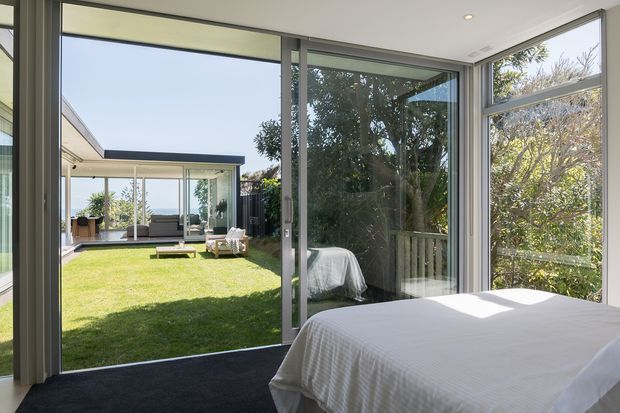 Bright contemporary bedroom with a white-covered bed positioned beside extensive floor-to-ceiling glazing and a large sliding glass door. The glazing opens directly onto a grassy courtyard or garden, with a covered outdoor seating area visible beyond. The room has dark carpet, slim grey window frames, white walls and ceiling, and abundant natural light.