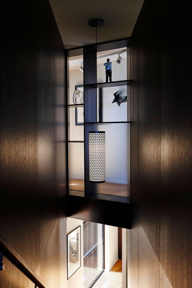 A dramatic narrow interior stair hall lined with dark timber wall panelling. A sculptural black metal and ribbed translucent-glass screen forms a mezzanine-like opening, with display niches, artwork and a pendant light. The lower level and stair are visible below, creating a strong vertical connection and layered views through the circulation space.