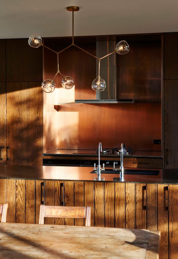 A warm, moody kitchen interior featuring dark timber cabinetry and vertically fluted timber panels on the island. A dark stone benchtop surrounds a polished stainless-steel sink and tapware, with a matching dark stone work surface and integrated cooktop at the rear. Copper-toned metal or reflective warm-toned panels form the splashback, while a sculptural multi-arm pendant with clear glass globes hangs above the kitchen. Strong natural sunlight creates dramatic highlights and shadows across the timber surfaces.