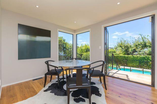 Bright, modern dining area with a round glass dining table and black dining chairs on a cowhide rug. Warm timber flooring, neutral painted walls, recessed ceiling lights, and extensive corner glazing create an airy connection to a timber deck and turquoise swimming pool outside.