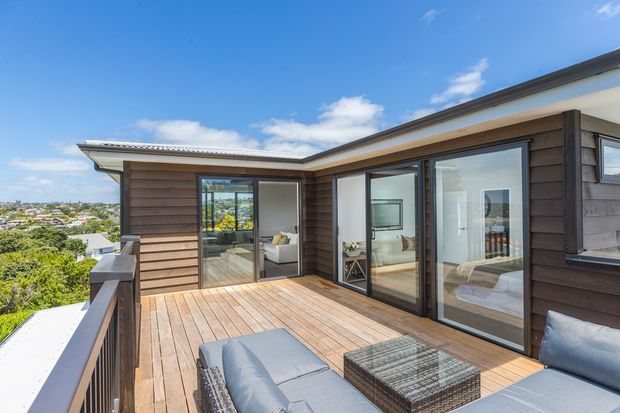 A contemporary upper-level timber-clad home with a spacious outdoor timber deck and dark-framed floor-to-ceiling sliding glass doors. The deck includes outdoor lounge furniture and overlooks a leafy suburban landscape beneath a bright blue sky. A bedroom and living area are visible through the glazing.