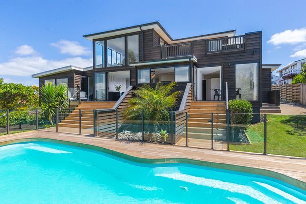 Contemporary multi-level home with dark horizontal timber cladding, extensive floor-to-ceiling glazing, balconies, and broad timber staircases leading to a poolside deck. The landscaped yard includes palms and manicured planting, with a bright blue swimming pool dominating the foreground.