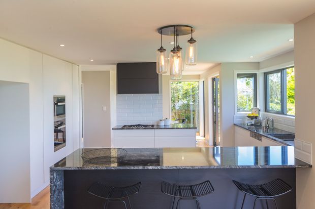 Bright contemporary kitchen with a large island and breakfast bar, dark stone benchtops, white cabinetry, integrated appliances, white subway-tile splashback, and pendant lighting. Windows and a glazed door provide views to greenery, while three black wire bar stools sit at the island.
