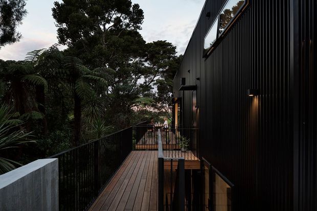 A narrow elevated timber walkway runs alongside a dark, vertically clad contemporary building, bordered by black metal balustrades. Dense mature greenery surrounds the structure, while large upper-level windows and warm exterior wall lights add contrast to the moody setting.