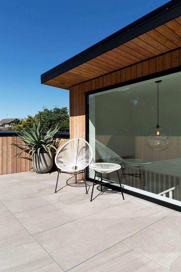 A sunny modern rooftop terrace or deck with large pale square paving tiles, timber-clad walls and soffit, black metal roof edging, and a full-height glazed sliding door. Outdoor wire-frame lounge chairs, a small table, and a large potted architectural plant create a relaxed seating area. The glazing reveals a contemporary interior with timber wall panelling, a dining table, and pendant lighting.