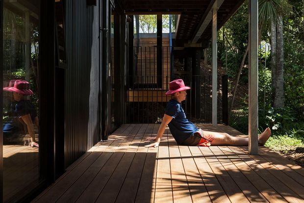 A shaded timber deck or covered outdoor walkway beside a dark-clad building, surrounded by lush vegetation. A seated person in a bright pink hat occupies the deck, while reflective glazing and vertical metal screens reveal the building edge and interior beyond. Strong sunlight creates linear shadows across the timber decking.