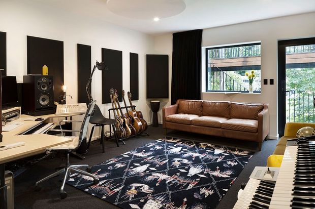 A stylish home music studio and lounge with a recording desk, studio monitors, multiple guitars, keyboards, a leather sofa, acoustic wall panels, and a bold patterned rug. Large windows bring in natural light and overlook greenery.