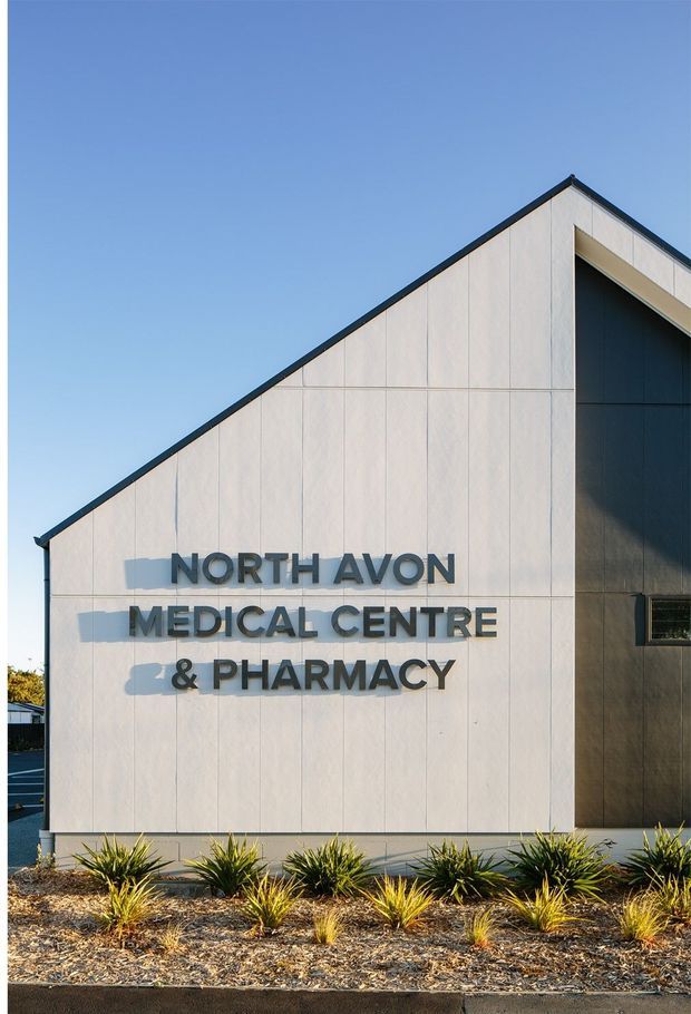 Exterior photograph of a contemporary medical centre and pharmacy with a tall, sharply pitched gable facade. The building is clad in pale vertically jointed panels with dark trim and a recessed dark entrance or window zone. Large raised lettering identifies the facility, while low ornamental plantings sit in a gravel bed along the frontage.