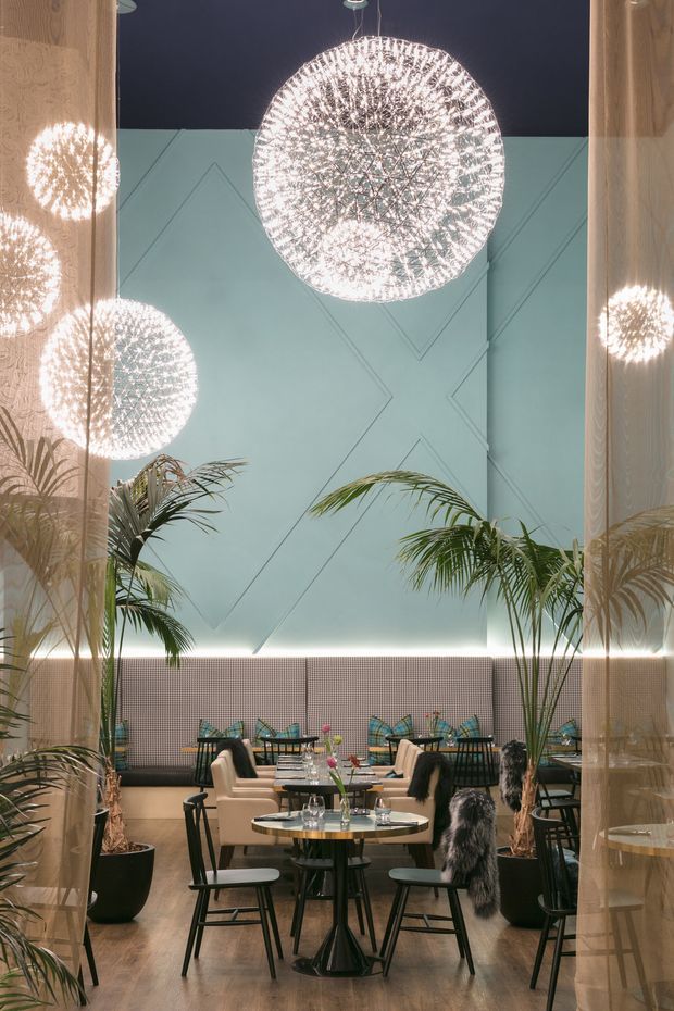 A stylish restaurant or café dining interior viewed through sheer curtains, featuring a geometric pale blue feature wall, warm timber flooring, patterned upholstered banquette seating, small round dining tables, black chairs, large potted palms, and dramatic spherical crystal pendant lights. The space has a refined, contemporary atmosphere with a strong decorative lighting focus.
