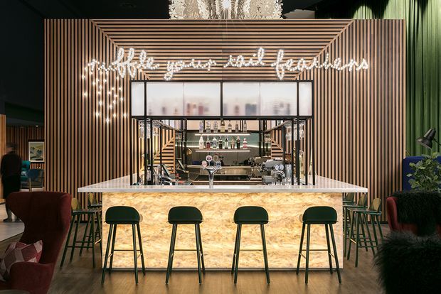 A sophisticated home bar or hospitality-style bar area with a luminous stone-fronted counter, four dark bar stools, a mirrored and glass-backed drinks display, and a dramatic illuminated script sign. Vertical timber battens frame the bar, while dark green walls, patterned wallpaper, lounge seating, and plants create a moody, luxe atmosphere.