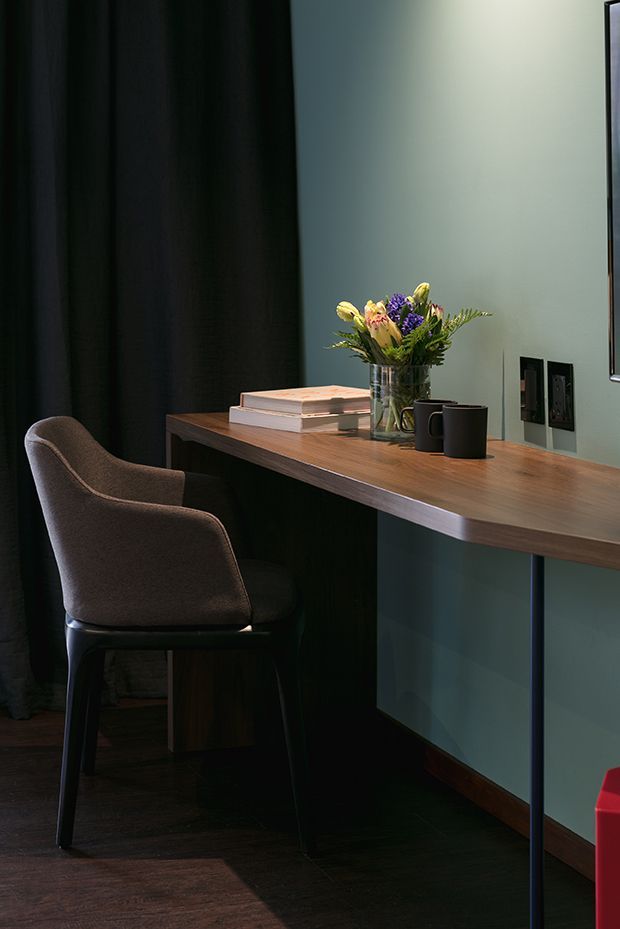 A moody, refined home workspace with a slim timber desk mounted against a muted blue-green wall, paired with a dark upholstered armchair. Black curtains, dark timber flooring, a vase of flowers, stacked books, ceramic mugs, and discreet wall outlets create a sophisticated study nook.