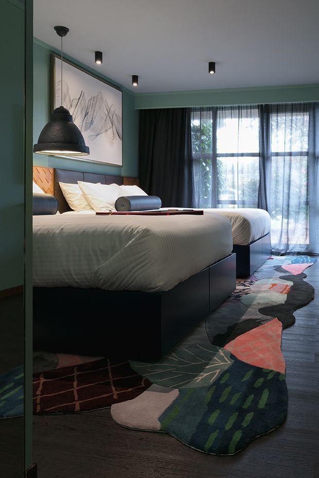 A moody, contemporary bedroom with two beds featuring dark built-in bases, white bedding, teal-green wall panelling, and a large abstract artwork above the headboards. Black pendant lights and recessed ceiling spotlights provide focused illumination, while sheer and blackout curtains cover a large window. A multicoloured organic-shaped patterned rug runs across the dark timber floor.