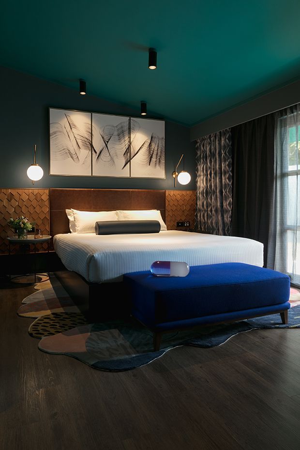 A moody, sophisticated bedroom with a large upholstered bed, dark teal walls and ceiling, warm wood flooring, patterned leather or wood wall panels behind the headboard, and dramatic layered curtains. Abstract monochrome artwork hangs above the bed, while pendant wall lights, a blue upholstered bench, and a sculptural patterned rug add bold decorative accents.