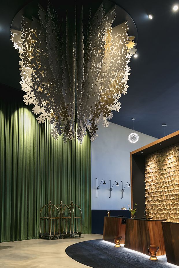 A dramatic, richly detailed reception lobby or commercial entry space. Floor-to-ceiling deep green curtains frame an oversized botanical metal pendant installation, while a sculptural textured stone or tile feature wall glows behind a dark timber reception desk. The room has a dark blue ceiling, polished light flooring, feature wall lighting, decorative wall lights, and a refined, theatrical atmosphere.