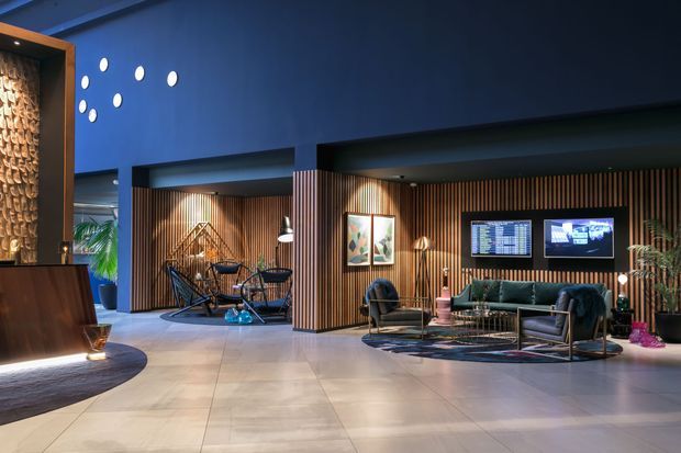 A spacious, dimly lit contemporary lobby or lounge with deep blue walls, pale large-format floor tiles, warm vertical timber-slat feature walls, and integrated display screens. The space includes a dark reception desk, lounge seating, coffee tables, decorative lighting, exercise bicycles in a recessed area, indoor plants, and patterned rugs. Dramatic spot lighting creates a refined, moody atmosphere.