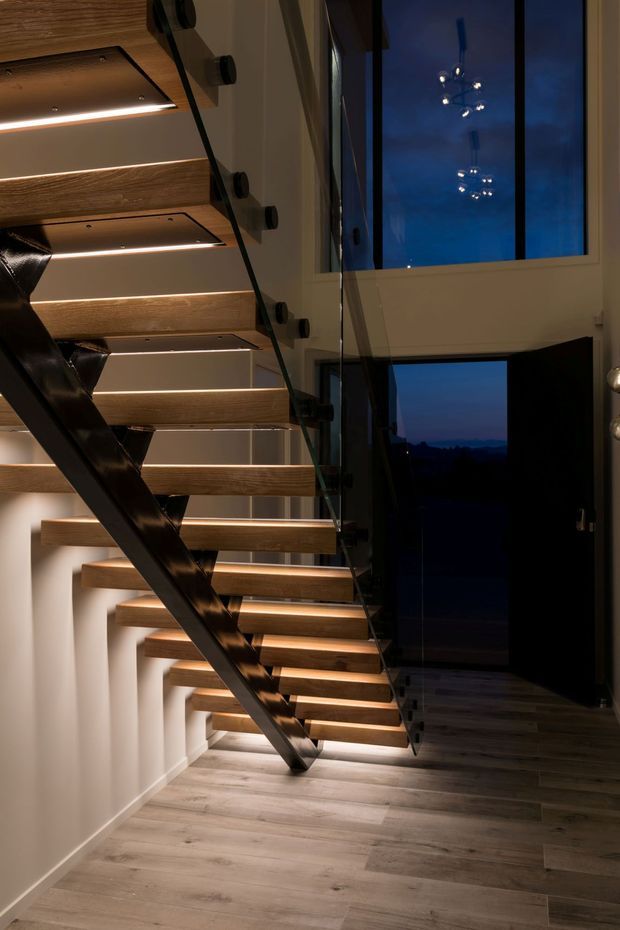 A dramatic modern stairway with floating timber treads, concealed warm LED lighting, a black steel diagonal stringer and clear glass balustrade. The stairwell has light neutral walls, timber-look flooring, tall glazing, and a dark entry door opening to a blue evening exterior. Reflections in the glass and glazing add depth to the compact vertical space.