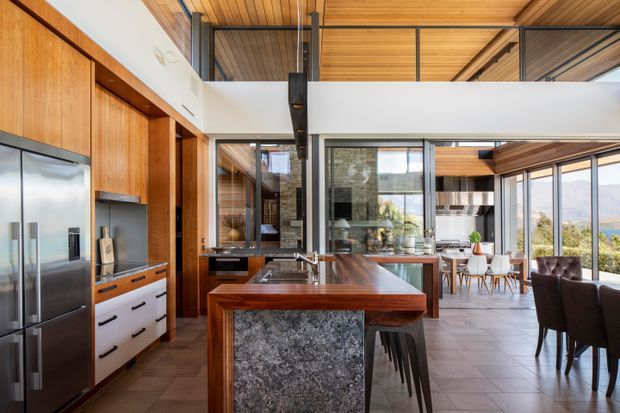 A bright, open-plan kitchen and dining area with a substantial timber island featuring a dark natural-stone waterfall end, integrated stainless-steel appliances, timber cabinetry, and a timber-lined ceiling. Black steel posts and framing support a mezzanine or upper-level void, while extensive glazing connects the dining area to the landscape beyond. A dining table and chairs are visible in the background, with bar stools at the island.