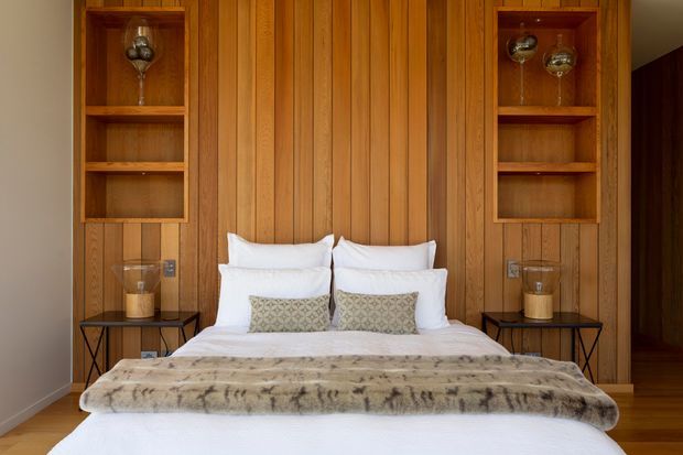 Warm, symmetrical bedroom with a centered bed against a full-height vertical timber-panel feature wall. Built-in timber shelving niches on both sides display glassware, with slim black bedside tables and lamps flanking the bed. The room has white bedding, patterned cushions, a plush neutral throw, timber flooring, and a calm, refined atmosphere.
