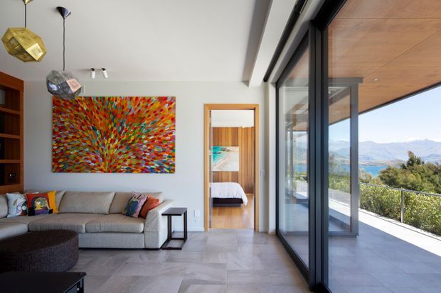 Contemporary living room with a large grey sectional sofa, colourful abstract artwork, sculptural pendant lights, and large-format grey floor tiles. Full-height black-framed glazing opens to a covered terrace with timber soffit and expansive mountain views. A bedroom is visible through the interior doorway.