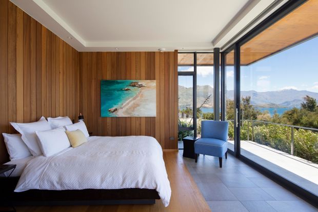 A modern bedroom with a large bed dressed in white linens, warm vertical timber wall panelling, and a blue accent chair beside floor-to-ceiling glazing. The glazing opens to a balcony or terrace with mountain and lake views, creating a bright, scenic retreat.