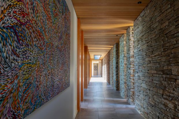 A long, refined architectural hallway with exposed stacked-stone walls on the right, warm timber ceiling and framing, pale large-format tiled flooring, and recessed lighting. A colourful abstract artwork occupies the foreground wall, while repeated openings and a skylight create depth and natural light toward the far end.