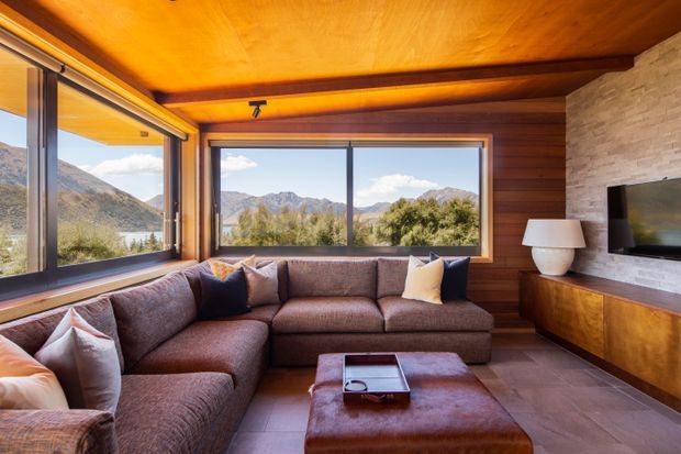 A warm, wood-lined living room with a large L-shaped sectional sofa, leather ottoman, built-in timber media cabinet, wall-mounted television, and expansive windows overlooking a mountainous landscape and lake. The room features a timber ceiling and walls, stone fireplace surround, and earthy, comfortable furnishings.