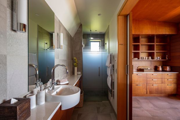 A refined bathroom viewed toward a frosted-glass shower enclosure, with twin vessel basins on a pale stone vanity, a large wall mirror, and warm timber cabinetry and shelving visible through an open doorway. Grey stone-look wall and floor finishes, brushed metal fittings, and soft natural light create a contemporary, calm interior.