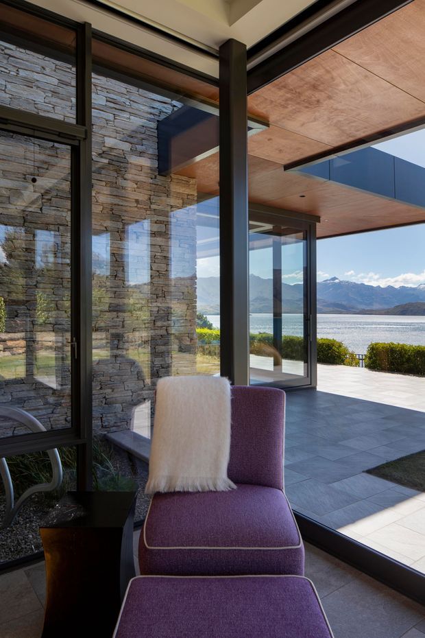 A contemporary interior seating area beside expansive dark-framed glazing, opening onto a paved terrace with sweeping views of a lake and mountainous landscape. The room features a stacked natural-stone wall, exposed timber soffit, dark structural columns and frames, and a purple upholstered lounge chair with ottoman.