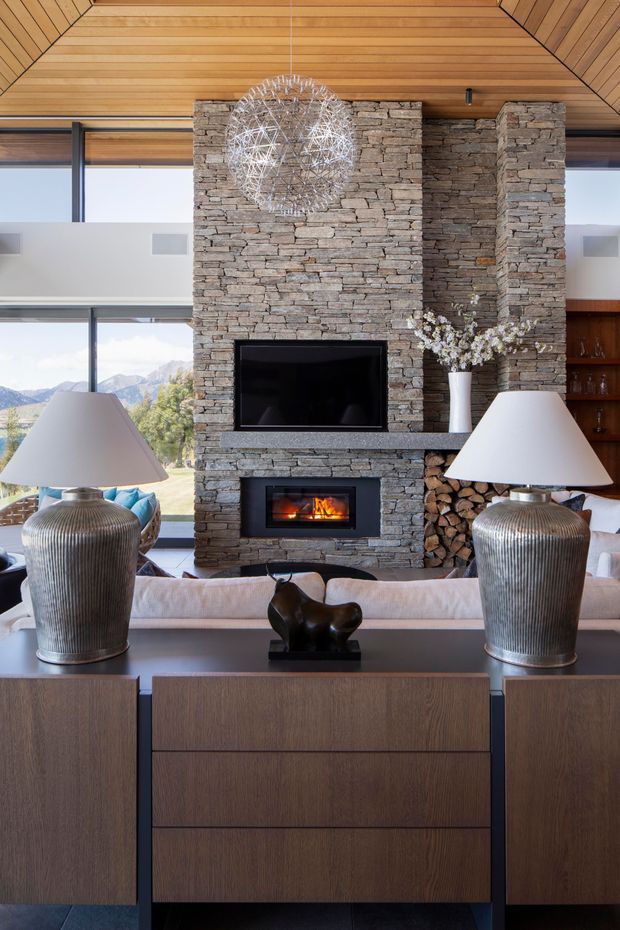 A refined living room with a central stacked-stone fireplace and integrated television, backed by expansive glazing overlooking a mountainous landscape. The room features a warm timber-lined ceiling, dark timber built-in cabinetry, contemporary lamps, a sculptural pendant, and neutral upholstered seating. Firewood is stored beside the fireplace, adding a rustic natural element to the otherwise polished interior.