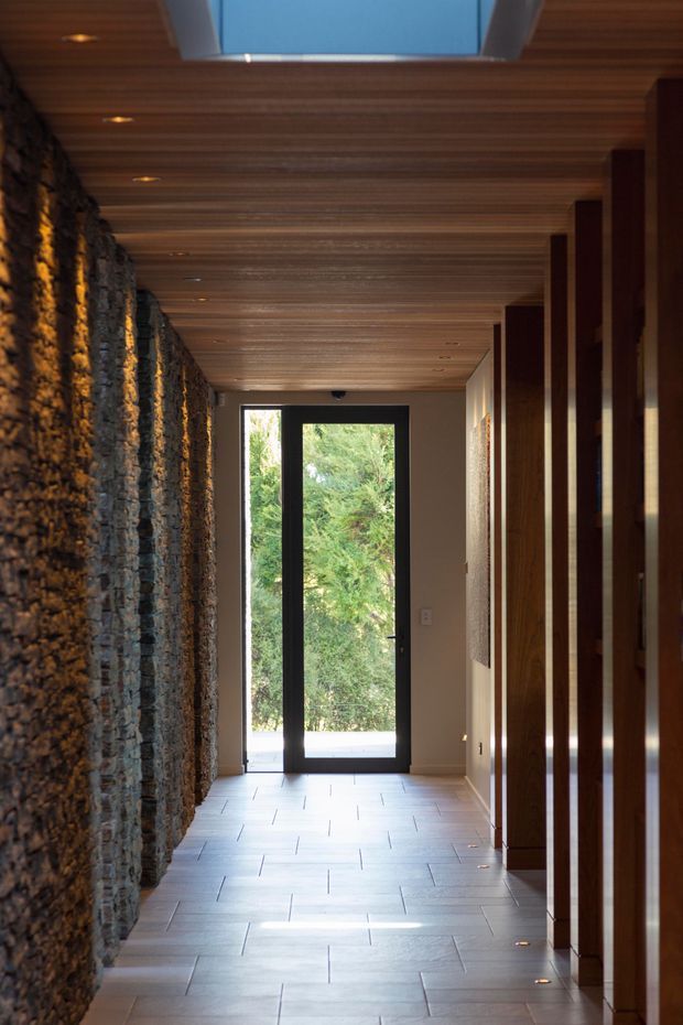 A narrow, refined entry hallway with a dramatic irregular stacked-stone feature wall on the left, warm horizontal timber ceiling lining with recessed spotlights and a large skylight, pale rectangular floor tiles, and dark timber vertical wall panelling and door frames on the right. A black-framed glazed door at the end opens toward greenery, bringing in natural light.
