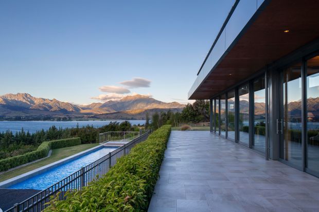 A contemporary lakeside residence with a long covered terrace, extensive floor-to-ceiling glazing, dark metal framing and a warm timber soffit. The terrace overlooks a narrow swimming pool, landscaped hedges, a lake and rugged mountains in the distance.