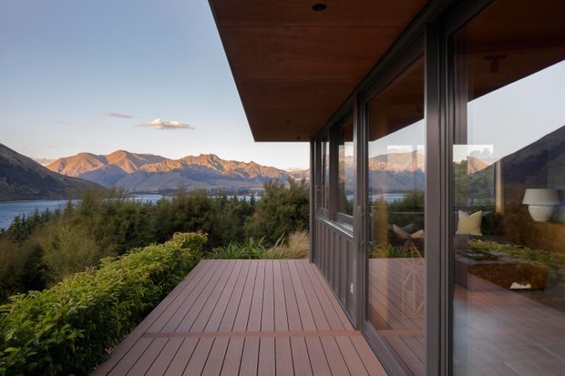 A contemporary lakeside home viewed from a timber deck, with extensive floor-to-ceiling glazing reflecting the surrounding mountain landscape. The scene shows a calm alpine setting, warm timber soffit and decking, dark window frames, landscaped planting, and a lake backed by rugged mountains.