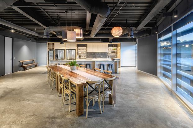 Open-plan commercial-style breakout and dining space with an industrial exposed-services ceiling, polished concrete floor, dark walls, large glazed windows, and a long timber communal table surrounded by mixed chairs and stools. A compact kitchenette and bar counter sit at the rear.