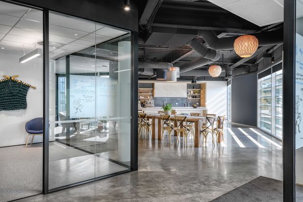 A bright contemporary commercial office interior with an open-plan breakout kitchen and dining area. The space features polished concrete flooring, exposed black ceiling services and ductwork, black-framed glass meeting-room partitions, timber cabinetry and shelving, a stone-look backsplash, pendant lighting, and large windows. A meeting room with a whiteboard is visible through the glass partition.