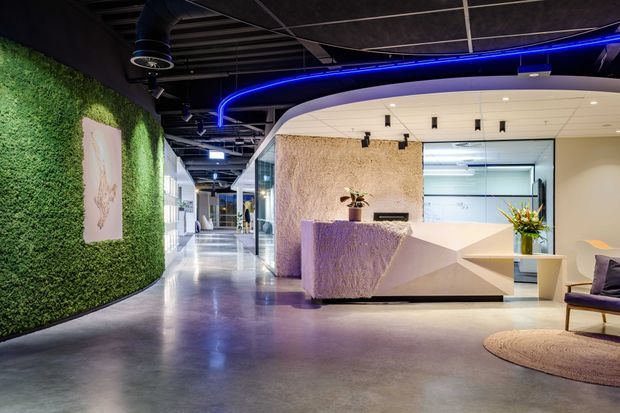 A contemporary commercial reception area with a sculptural white reception desk, textured stone-like feature wall, glass-partitioned offices, polished concrete flooring, and a curved living green wall. Exposed dark ceiling services and vivid blue linear lighting create a bold, high-tech atmosphere.