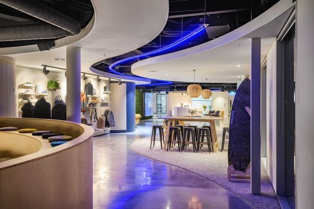 A contemporary commercial showroom or retail interior with an open-plan layout, polished concrete flooring, curved built-in seating, curved sculptural ceiling forms, exposed black ductwork, recessed display shelving, clothing racks, and a central communal table with bar stools. Blue linear lighting adds a dramatic accent to the otherwise neutral and warm timber-toned fit-out.