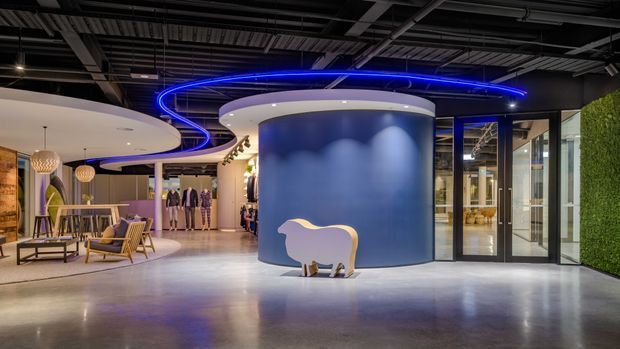 A sophisticated commercial interior with an open reception or lounge area, polished concrete flooring, a large curved blue feature wall, glass entry doors, sculptural ceiling forms, and dramatic blue LED lighting. Lounge seating, display mannequins, decorative lighting, and a green wall contribute to a contemporary showroom or retail environment.