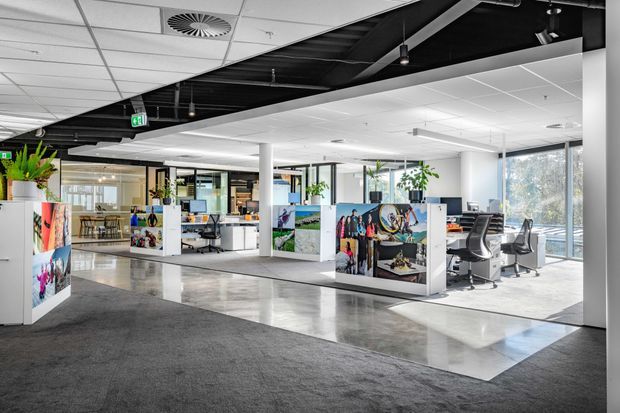 Bright open-plan commercial office interior with workstations, ergonomic chairs, partition walls displaying colourful graphics, extensive glazing, polished concrete flooring, carpeted circulation areas, and a partially exposed black ceiling with suspended lighting and services. Indoor plants and abundant daylight create a contemporary workplace atmosphere.