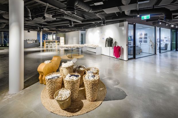 A spacious contemporary retail or showroom interior with polished concrete flooring, exposed black services and ductwork, white structural columns, glass-partitioned rooms, and minimalist display fixtures. Woven baskets and sculptural furniture form a central product display, while clothing and framed items are presented around the perimeter.