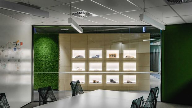 Contemporary commercial showroom or office interior with a long meeting table and black mesh chairs in the foreground. A backlit display wall with recessed niches showcases products or objects, flanked by a vertical green wall and glass partitions. The suspended grid ceiling, exposed services, linear pendant lighting, and polished dark floor create a clean, modern workplace atmosphere.