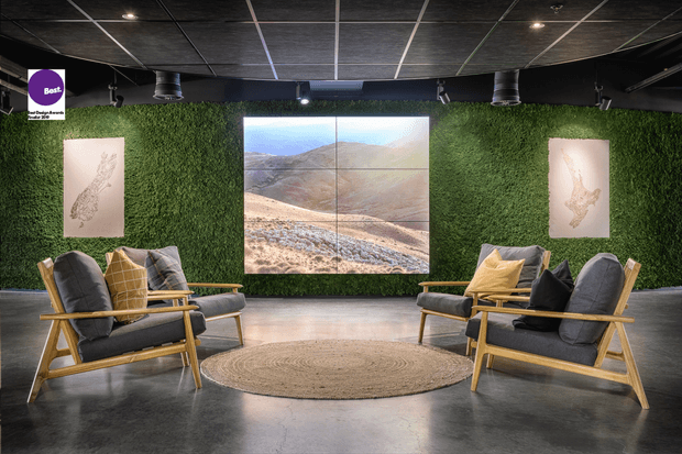 A dark, refined lounge or reception area with moss-green living walls, exposed black ceiling grid and track lighting, polished concrete flooring, and paired timber-framed lounge chairs arranged around a round woven rug. A large multi-panel landscape image forms the focal point, with minimalist artwork mounted on either side.